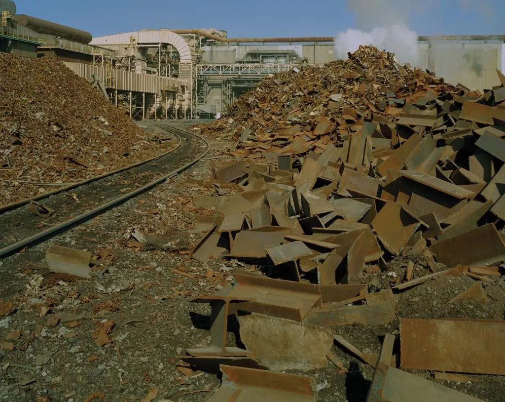 In Nucor-Yamato's scrap yard, piles of ferrous scrap, including discarded ends of beams and finely shredded metal fragments, lie ready for transporting into the plant, where they will be melted into liquid steel. Behind them stands the air-handling equipment that removes and treats hot, dusty exhaust from the melting operations.