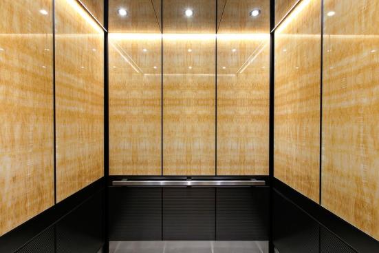 LEVELe-105 Elevator Interior with upper panels in ViviStone Honey Onyx, Standard finish; lower panels in Fused Graphite, Sandstone finish, Dallas pattern; Handrail panel in Fused Graphite, Seastone finish; Quadrant Handrail in Satin Stainless Steel; Elevator Ceiling in Fused Nickel Silver, Mirror finish at 176 Broadway, New York, New York