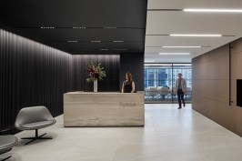 The Tiptree reception desk of solid travertine stone anchors the entry, set against the background of a black wood panel grille. The separation of executive areas from collaborative common areas is visually represented by the division of light and dark woods.