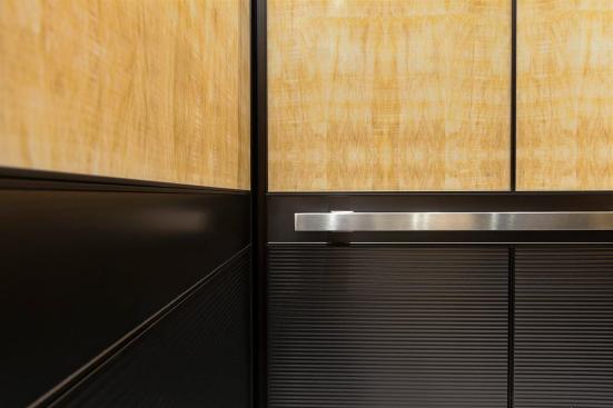 LEVELe-105 Elevator Interior with upper panels in ViviStone Honey Onyx, Standard finish; lower panels in Fused Graphite, Sandstone finish, Dallas pattern; Handrail panel in Fused Graphite, Seastone finish; Quadrant Handrail in Satin Stainless Steel at 176 Broadway, New York, New York