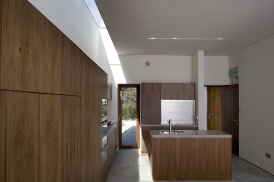 A wall of walnut veneered built?in-furniture runs the full length of the new living block beneath a rooflight, incorporating the stove, desk and kitchen in one unified element and focusing the occupant?s gaze towards the views to either end.