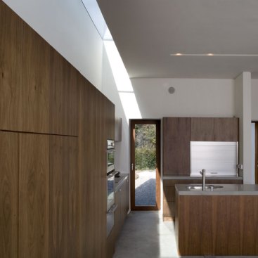 A+wall+of+walnut+veneered+built%e2%80%93in-furniture+runs+the+full+length+of+the+new+living+block+beneath+a+rooflight%2c+incorporating+the+stove%2c+desk+and+kitchen+in+one+unified+element+and+focusing+the+occupant%e2%80%99s+gaze+towards+the+views+to+either+end