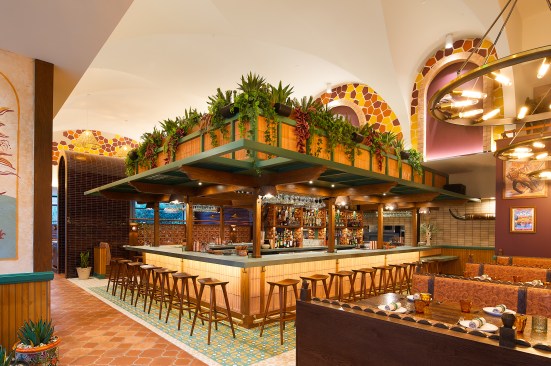 The middle of the main dining room is home to the lower-level bar, featuring overhead shutters and raised booths with custom light fixtures carved from old, knotted wood. 