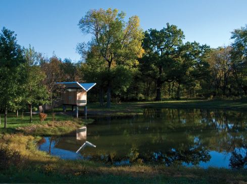 Meditation Hut III "Victor"- Champaign, IL The owners of a forested property wanted a quiet space to observe nature. Entry is across a visually kinetic approach ramp and through an obscured door detailed like the cedar walls. Inside an oversized window creates a composed view of mature trees. Adjacent to this is a miniature tea cabinet. A raised platform in the main space supports three tatami mats. The location along the north pond edge allowed the development of several effluvial sensations. Throughout the day water reflections are projected onto the soffit. The roof channels rainwater to a central spout over the pond. A horizontal window in the tatami room frames a meditative fragment of water. The floor of glossy ebonized birch has the sensation of a deep still pool--the grass tatami mats become an island within an island. The result is an interior volume that is protective and serene but alive with subtle energy. 
Architect: Jeffery S. Poss Architect